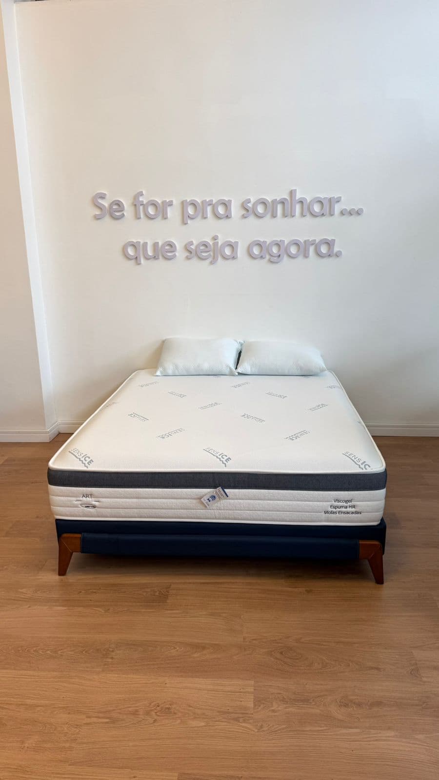 Colchão branco com travesseiros sobre base azul sob frase motivacional na parede branca.