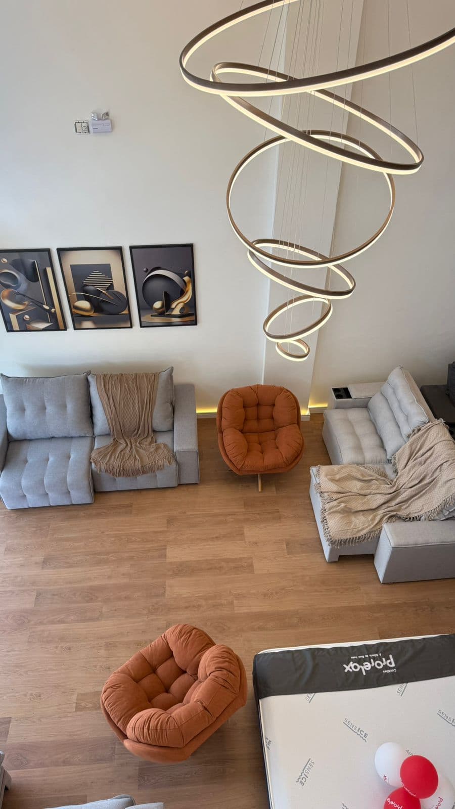 Sala moderna com sofás cinza, poltronas laranjas, quadros abstratos e um lustre pendente em espiral.